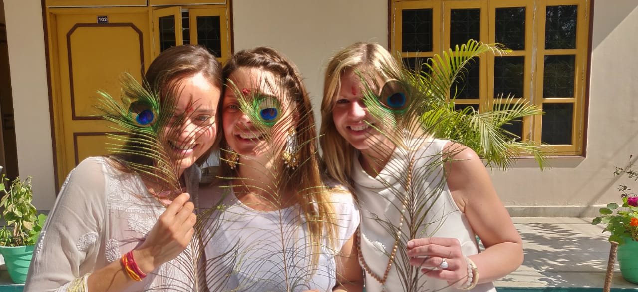 Three smiling students hold peacock feathers, foreheads marked with tilak, at the Yoga Bhawna Mission campus in Uttarkashi.