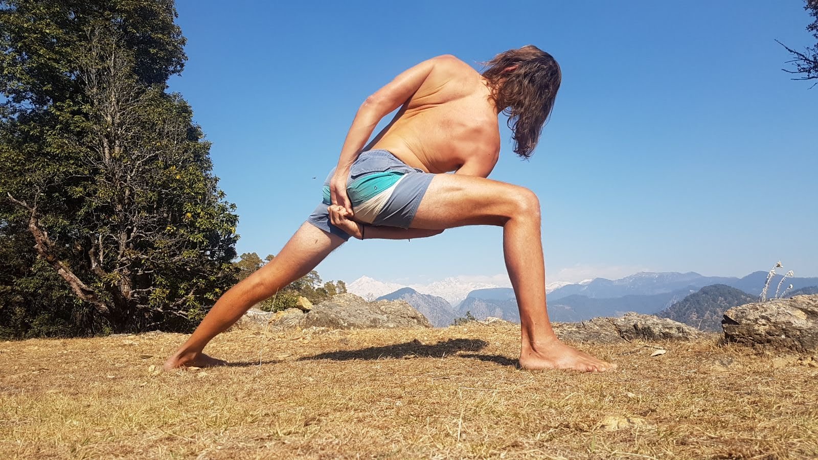 A yoga student holds a revolved side-angle lunge on a Himalayan hilltop, snow-capped peaks behind, during a Yoga Bhawna Mission course in Uttarkashi.