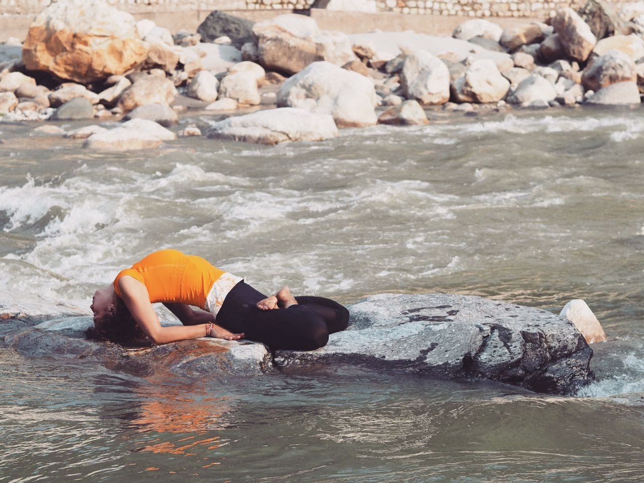 A yoga practitioner rests in a supported backbend on a boulder in the flowing Bhagirathi river at Uttarkashi.