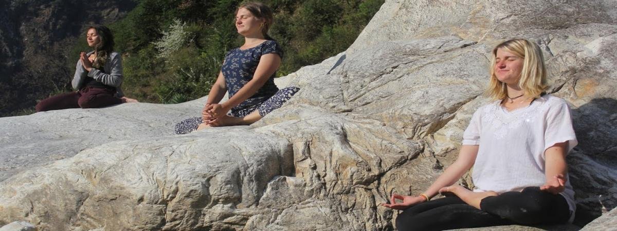 Three students meditate in lotus pose on sunlit rocks beside the river during a Yoga Bhawna Mission retreat in Uttarkashi.