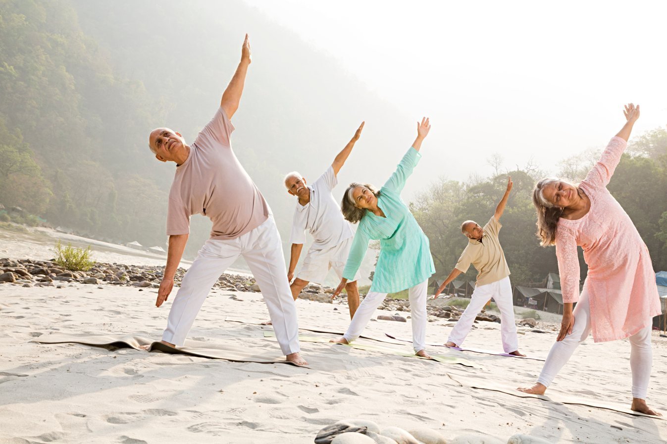 A group of older students practise trikonasana, triangle pose, together on the sand beside the river with the Himalayan foothills behind them.