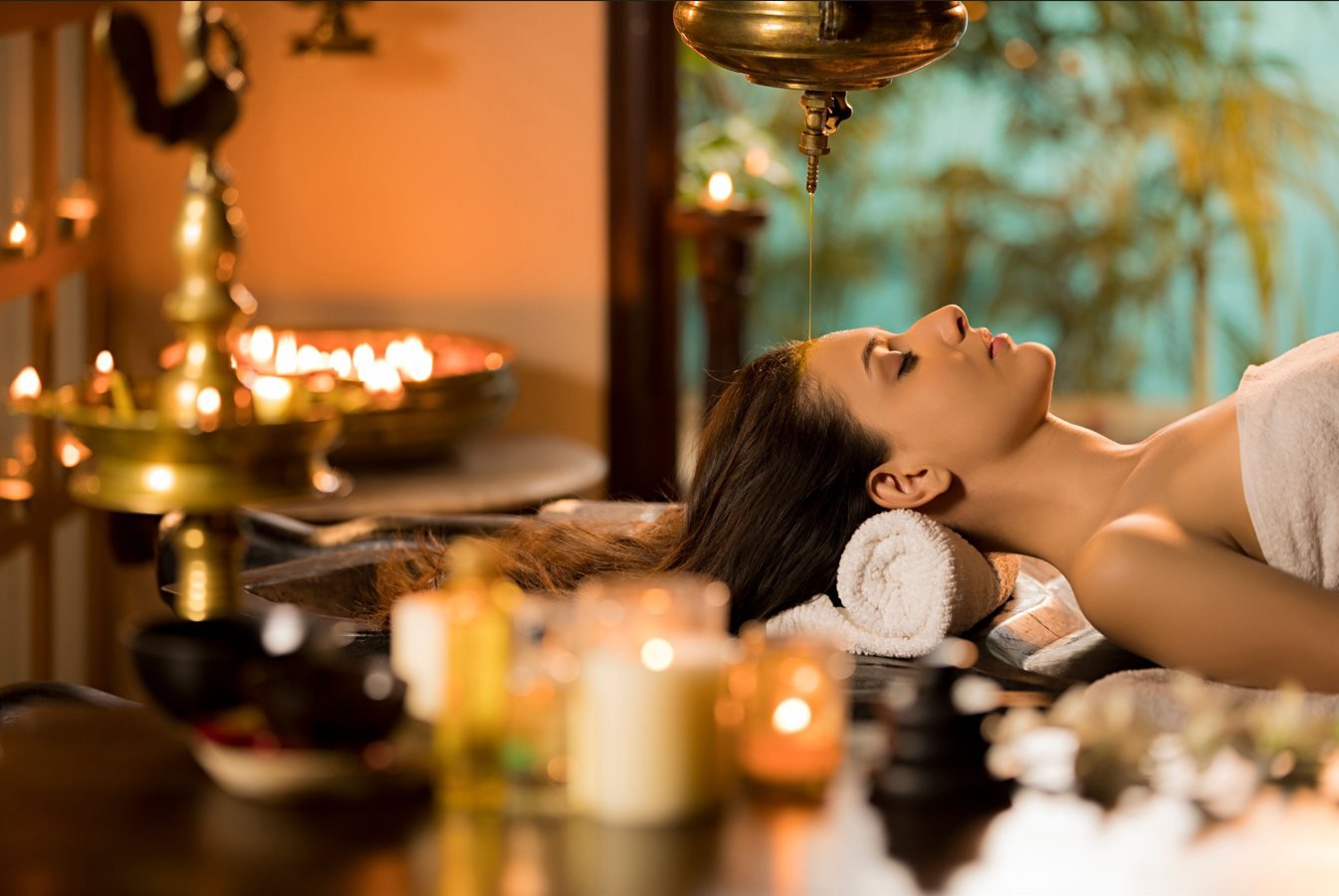 An Ayurvedic shirodhara treatment in progress, with warm oil poured in a steady stream onto the forehead in a candlelit therapy room.