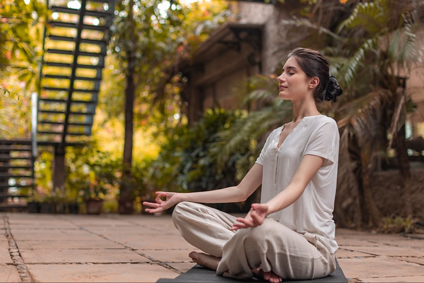 A woman meditates cross-legged on a stone courtyard among trees in the late afternoon light.