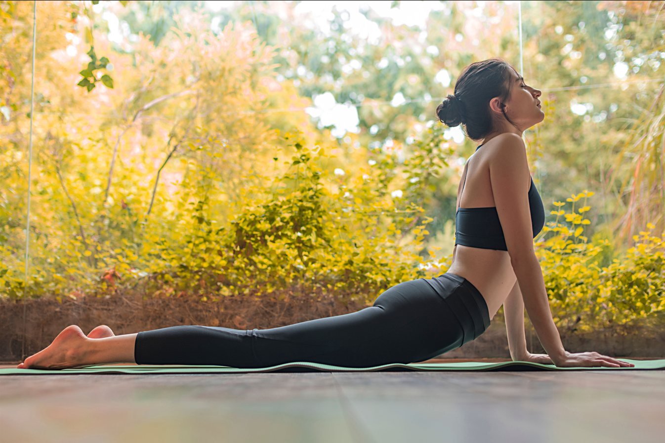 A student moves through upward-facing dog on a mat outdoors with autumn trees behind.